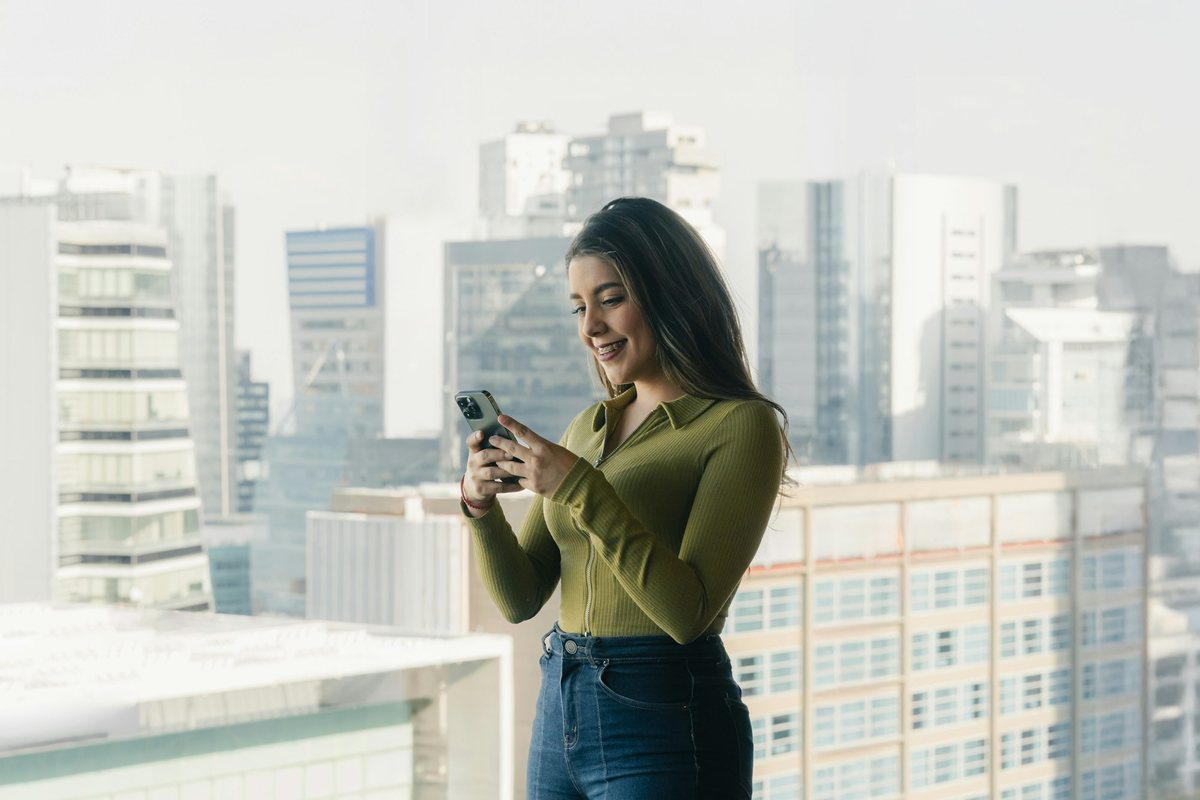 Residente sonríe mientras revisa su teléfono junto a la ventana de su departamento, con torres al fondo